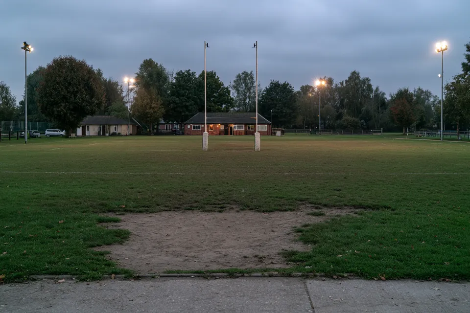 Terrain d’un club de rugby local au crépuscule, poteaux en H et petit club-house en arrière-plan